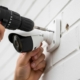 Close-up of a person installing an outdoor security camera on a white brick wall using a power drill, with ‘American Alarm’ in the bottom right corner.