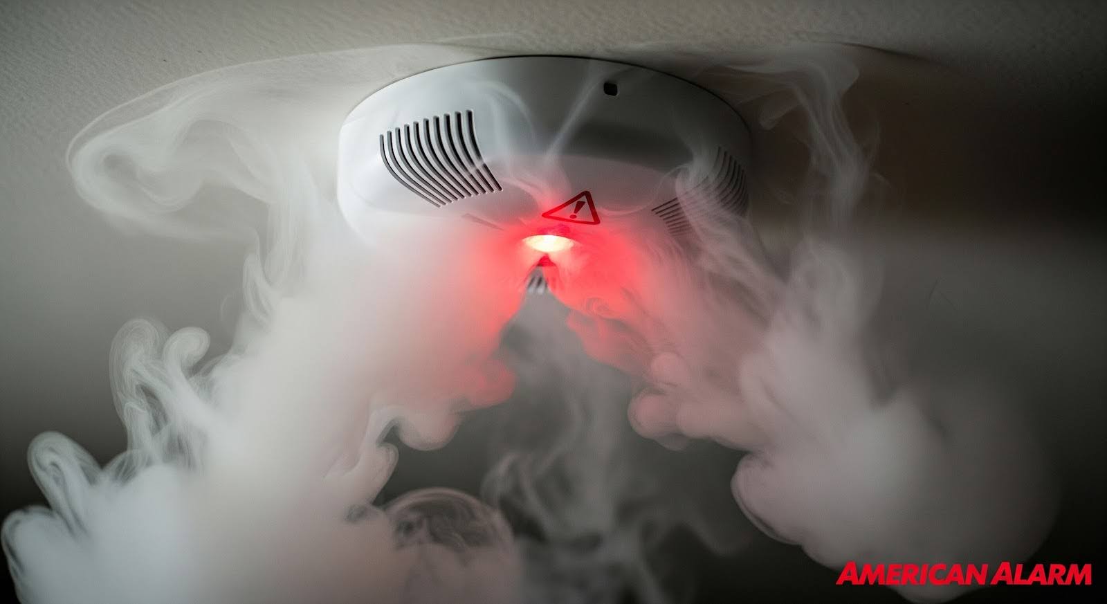 A smoke detector emitting a red warning light as thick smoke surrounds it, with ‘American Alarm’ in the bottom right corner.