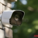 A wall-mounted outdoor security camera in focus against a blurred green background, with ‘American Alarm’ in the bottom right corner.