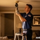 A technician on a ladder replaces a ceiling-mounted smoke detector in a warmly lit living room, with ‘American Alarm’ shown in the bottom right corner.