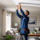 A technician on a ladder installs a smoke detector in a bright living room, with ‘AMERICAN ALARM’ shown in the bottom right corner.